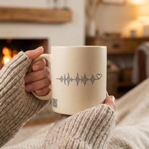 May include: Cream ceramic mug with a black sound wave and heart design. The mug is held by a person wearing a grey sweater. A QR code is visible on the mug. The background shows a fireplace and a cosy living room setting.