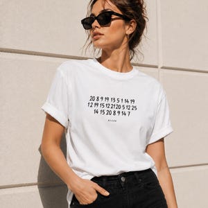 Op de afbeelding: Een wit T-shirt met korte mouwen en een zwarte grafische print van drie rijen cijfers en de tekst AIZ26 in het midden op de borst. Het shirt heeft een ronde hals, opgerolde mouwen, een zwarte broek en een zwarte zonnebril.