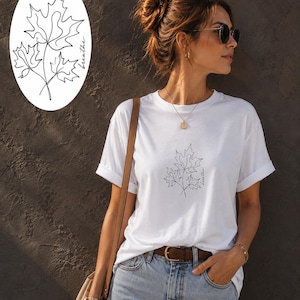 Peut inclure: T-shirt blanc avec un dessin minimaliste de feuilles d'érable et le mot "breathe". Le motif est centré sur le t-shirt. Le modèle porte un jean bleu et une ceinture marron.