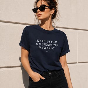 May include: A dark navy blue short-sleeved t-shirt featuring a white graphic print of three rows of numbers and the text A1Z26. The shirt has a crew neckline and rolled sleeves, paired with black denim jeans and black sunglasses.