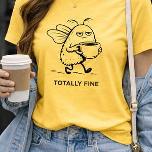 May include: Yellow t-shirt with a black cartoon character holding a coffee cup, the text "TOTALLY FINE" is below the character. The person is wearing blue jeans and a denim jacket. A person is holding a coffee cup.