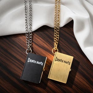 May include: Two pendant necklaces featuring miniature book charms. One charm is black with silver chain, the other is gold with a gold chain. Both charms read "Death Note" in white lettering. The background is a dark wood surface.