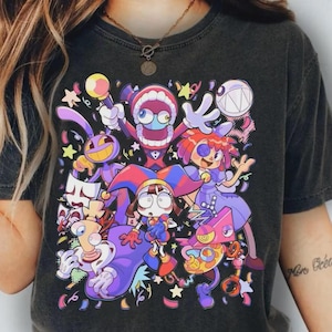 May include: A charcoal gray t-shirt featuring a vibrant, multi-colored graphic print of various cartoon characters in a festive setting with confetti and stars. The design includes a central jester figure, a toothy creature, and other whimsical shapes.