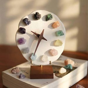 May include: A unique clock featuring a white, irregular-shaped face with colorful gemstones marking the hours. The clock has dark brown wooden hands and a matching wooden base. The clock is displayed on a book, with additional gemstones scattered around.