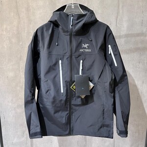 May include: A charcoal grey Arc'teryx hooded jacket featuring a full-length black front zip, white zipped chest and sleeve pockets, and a white brand logo on the chest. The jacket includes attached product tags and a Gore-Tex branding label.