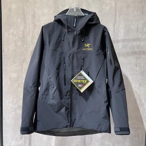 May include: A black hooded jacket featuring a full-length front zip, two vertical chest pockets, and a yellow Arc'teryx logo on the upper left chest. A Gore-Tex tag is attached to the zip, and the cuffs have adjustable hook-and-loop straps.
