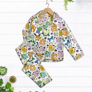 May include: White pajama set with a colorful pattern of various cartoon characters. The long-sleeved top has a collared neckline and button closure. The matching pants complete the two-piece sleepwear set.