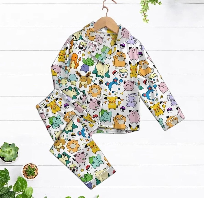 May include: White pajama set with a colorful pattern of various cartoon characters. The long-sleeved top has a collared neckline and button closure. The matching pants complete the two-piece sleepwear set.