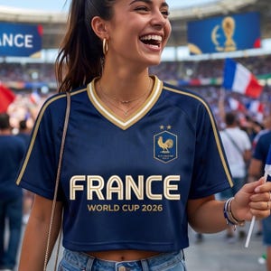 May include: A navy blue crop top jersey with gold trim and the word "FRANCE" in gold lettering. The jersey has a V-neck and features the French rooster emblem and the text "WORLD CUP 2026". The woman is smiling and wearing gold hoop earrings.