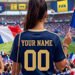 May include: Navy blue soccer jersey with gold lettering and trim. The jersey features the text "YOUR NAME" and the number "00". The background shows a stadium with spectators and flags.