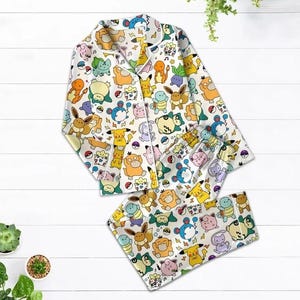 May include: White pajama set featuring a colorful pattern of various cartoon characters. The long-sleeved top has a button-down front and a notched collar. The matching pants have an elastic waistband. The set is made of a soft, lightweight fabric.