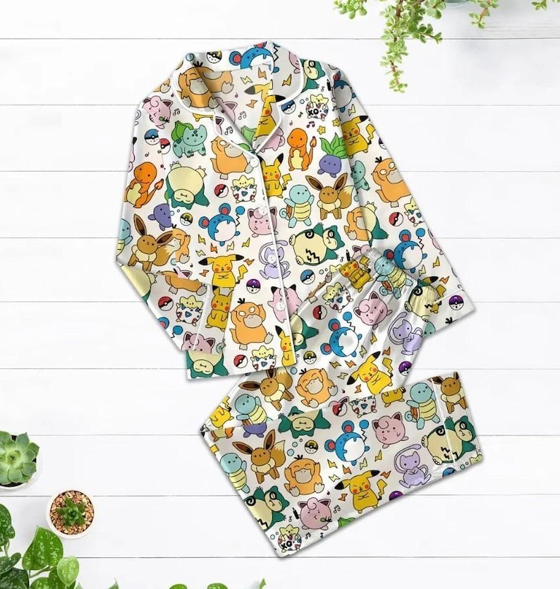 May include: White pajama set featuring a colorful pattern of various cartoon characters. The long-sleeved top has a button-down front and a notched collar. The matching pants have an elastic waistband. The set is made of a soft, lightweight fabric.