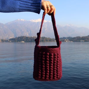 May include: A burgundy crochet bucket bag with a textured, bobble stitch design. The bag has a matching strap and is held up against a scenic lake and mountain backdrop. The bag is a handmade accessory.