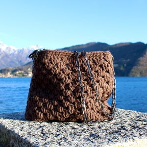 May include: A brown, crocheted handbag with a silver chain strap rests on a stone surface. The bag is square-shaped and has a textured pattern. The background features a lake, mountains, and a clear blue sky.