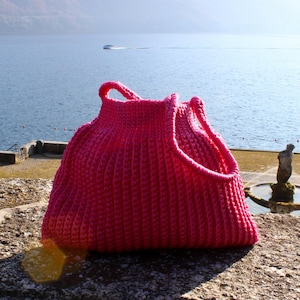 May include: A vibrant pink, handmade crochet tote bag with a structured design. The bag features a rounded shape with two handles and a textured stitch pattern. The bag is sitting on a stone surface with a scenic background.