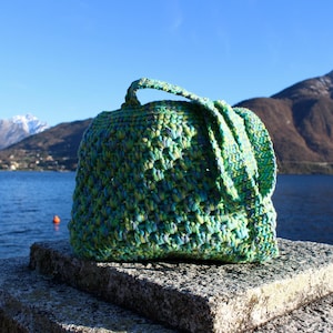 May include: A handmade crochet bag in shades of green and blue sits on a stone surface. The bag has a handle and a flap. The background features a lake, mountains, and a clear blue sky.
