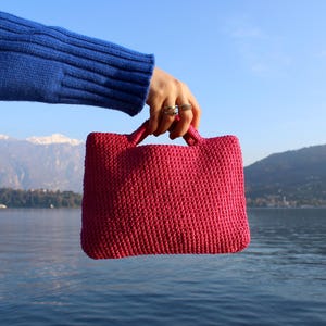May include: A vibrant fuchsia-colored tote bag with two handles is held up against a backdrop of a lake and mountains. The bag has a textured, woven appearance. The person holding the bag is wearing a blue sweater.
