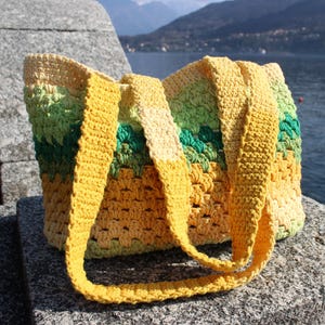 May include: A handmade crochet tote bag in shades of yellow, green, and cream. The bag features long, sturdy straps and a textured stitch pattern. The bag is sitting on a stone surface with a lake and mountains in the background.