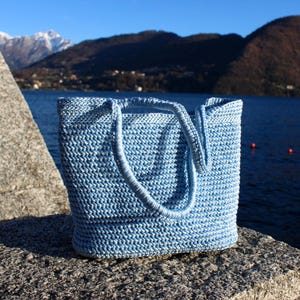 May include: A light blue, hand-crocheted tote bag with two handles. The bag is made of a textured yarn and sits on a stone surface. The background features a lake, mountains, and a clear blue sky.