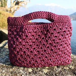 May include: A handmade, burgundy crochet tote bag with a sturdy handle. The bag has a textured, open weave pattern and is sitting on a stone surface. The background features a lake and mountains.