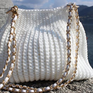 May include: A white, textured crochet handbag with a gold chain strap. The bag is sitting on a stone surface, with a body of water and mountains in the background. The chain is gold with white leather woven through it.