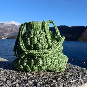 May include: A green, handmade crochet bag with a drawstring closure and two handles. The bag is sitting on a stone surface with a lake and mountains in the background. The bag's texture is visible.