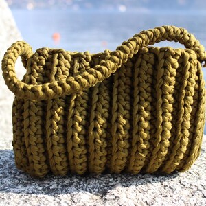 May include: Olive green crochet handbag with a braided handle. The handmade bag has a textured, ribbed design and is sitting on a stone surface. The bag is a stylish accessory for carrying essentials.