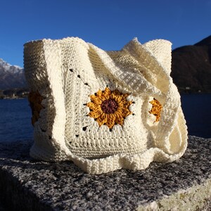 May include: Cream-colored crochet tote bag featuring a large sunflower design with yellow petals and a dark maroon center. The bag is square-shaped with a ruffled edge and a strap. The background includes a lake, mountains, and a clear blue sky.