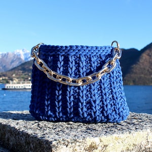 May include: A cobalt blue crochet handbag with a gold-tone chain strap. The bag is sitting on a stone surface with a lake and mountains in the background. The bag has a boxy shape.