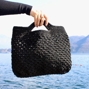 May include: A black crocheted tote bag with two handles is held up against a backdrop of a lake and mountains. The bag has a textured, open weave design. The bag is a solid black color.