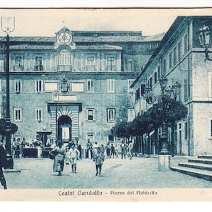 May include: A vintage postcard depicting a street scene in Castel Gandolfo, Italy. The image shows a large building with a clock tower and arched entrance, with people in the square. The text "Castel Gandolfo - Piazza del Plebiscito" is visible.