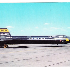 Könnte beinhalten: Ein schwarzes NASA X-15 Raketenflugzeug auf einer Landebahn, mit dem Text "U.S. AIR FORCE" und der Nummer "66671" sichtbar. Das Flugzeug hat gelbe Akzente an Heck und Flügeln, vor blauem Himmel.