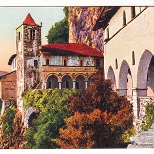 May include: Vintage postcard showing a building complex on a cliffside. The buildings have red tile roofs and arched walkways. The background features greenery and a body of water. A stone staircase leads up.