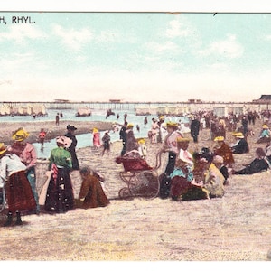 May include: Vintage postcard titled "THE BEACH, RHYL." depicting a crowded beach scene. People in period clothing are gathered on the sand, with a pier and buildings in the background. The image has a faded, aged appearance.