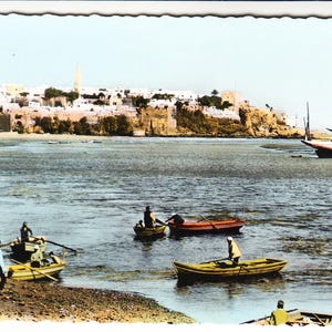 May include: Vintage postcard depicting a coastal scene with several small boats on the water. A sailboat is beached in the distance, and a city with white buildings and a tower is visible on the horizon. The water is calm, and the sky is a pale blue.