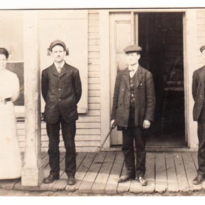 Puede incluir: Fotografía vintage en blanco y negro que muestra a cuatro personas frente a un edificio. Una mujer con un vestido largo está a la izquierda, mientras que tres hombres con trajes y sombreros están a la derecha. La imagen tiene un tono sepia.