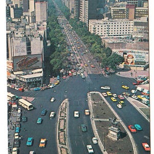 May include: A vintage postcard showing a wide city street with tall buildings and green trees. Numerous cars and buses travel on the multi-lane road. A large billboard is visible on the left side of the image.