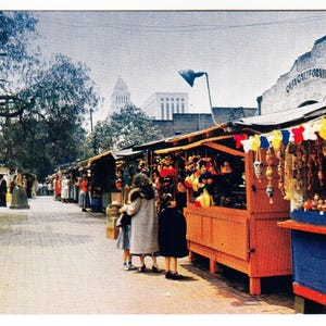 May include: Vintage postcard depicting a bustling outdoor market scene. Stalls with colorful displays line a brick-paved street. People browse the merchandise. The building in the background has the words "Casa California".