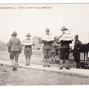 May include: Vintage black and white postcard titled "War Bread, Fort Logan, Colorado." Several uniformed soldiers carry stacks of bread. A horse-drawn cart and a pile of supplies are visible in the background.