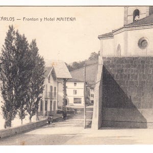 May include: Vintage black and white postcard featuring a street scene in Valcarlos, Spain. The postcard shows buildings, trees, and a church with a bell tower. The text "5 VALCARLOS - Fronton y Hotel MAITENA" is visible at the top.