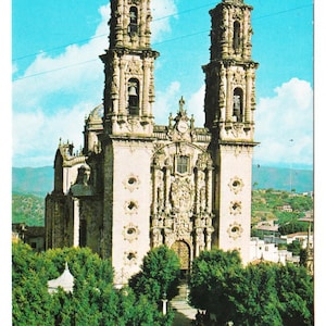 May include: Vintage postcard featuring a detailed, ornate church with two bell towers under a blue sky. The beige stone facade is intricately carved. Green trees and a glimpse of a town are visible in the foreground.