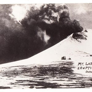 May include: Vintage black and white postcard depicting the Mt. Lassen eruption on June 14, 1914. The image shows a snow-covered mountain with a large plume of dark smoke billowing from the summit. The text "Mt. Lassen Eruption, June 14, 1914" is visible.