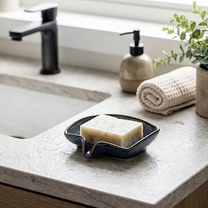 May include: A square, dark blue ceramic soap dish with a small spout, holding a rectangular bar of off-white, speckled soap. The dish features a glossy finish with a dark rim, designed to hold a single bar of soap securely on a countertop.