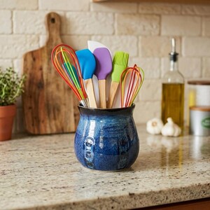May include: A deep blue ceramic utensil crock holds a collection of kitchen tools with wooden handles, including a red and green wire whisk, a blue silicone spatula, a purple silicone spatula, a green silicone brush, and an orange wire whisk.