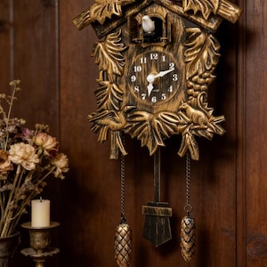 May include: A decorative cuckoo clock with a gold finish, featuring a bird on top and a bird emerging from a small door. The clock has a traditional design with a pendulum and weights, and is mounted on a wooden wall.