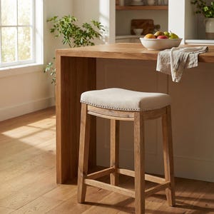 May include: A wooden bar stool with a light beige upholstered seat and a curved back. The stool has a sturdy, four-legged design with a footrest and is made of light-colored wood. The seat is trimmed with small, decorative nailheads. The stool is in a kitchen setting.