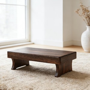 May include: A dark brown wooden step stool with a rectangular top and curved legs. The stool is placed on a cream-colored, shaggy rug. A white vase with dried flowers sits in the background near a window.