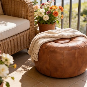 May include: A brown leather ottoman with a cream-colored throw blanket on a balcony. The ottoman is round and has stitched detailing. A wicker sofa and potted flowers are also visible, creating a cozy outdoor space.