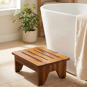 May include: A square, wooden step stool with a rich brown finish. The stool has four legs and three evenly spaced slots cut into the top. It is designed for use in a bathroom setting.