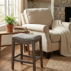 May include: A gray upholstered bar stool with a curved seat and nailhead trim. The stool has a dark wood frame and is positioned in front of a beige armchair with a blanket draped over it. A small table with a potted plant and books is nearby.